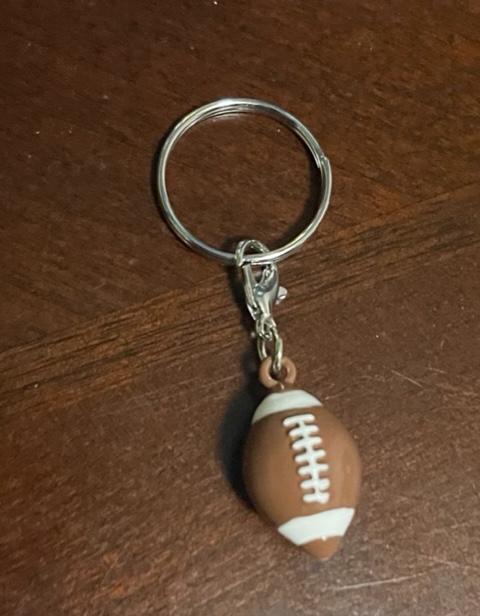 Small Brown and White Football Charm hanging from a silver chain, laying on top of a wooden table.