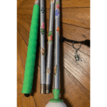 A foldable mobility cane with silver segments decorated with tropical flower and leaf stickers. It has a green rubber grip and a green rolling ball tip. A frog charm keychain is attached to the elastic cord.