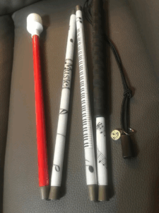 A musical-themed cane featuring segments wrapped in sheet music graphics, piano keys, and musical notes. The handle is black, and the tip is white with a red shaft segment leading into it. A small charm with a music note is attached.