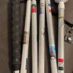 A white cane adorned with mini flags and landmark photos from around the world, including the Eiffel Tower and Leaning Tower of Pisa. It has a black grip and a white marshmallow-style tip. A tiny camera charm is attached.