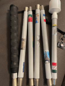 A white cane adorned with mini flags and landmark photos from around the world, including the Eiffel Tower and Leaning Tower of Pisa. It has a black grip and a white marshmallow-style tip. A tiny camera charm is attached.
